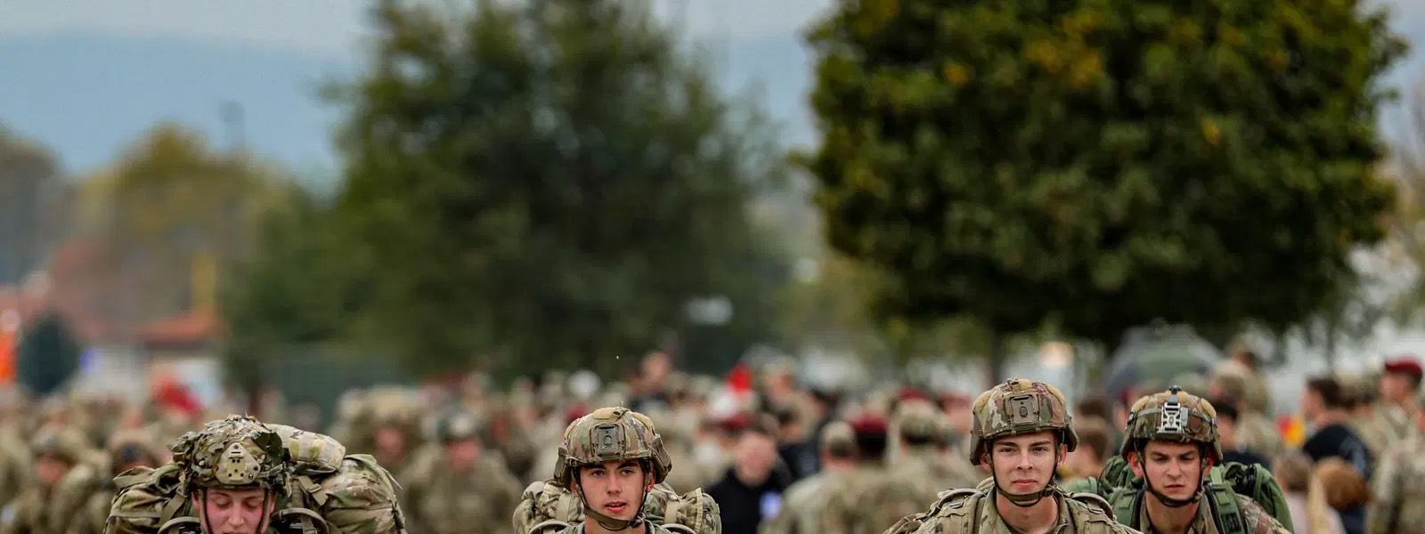 Soldiers carrying heavy rucksacks walk towards the camera during an endurance march, with a large group behind them